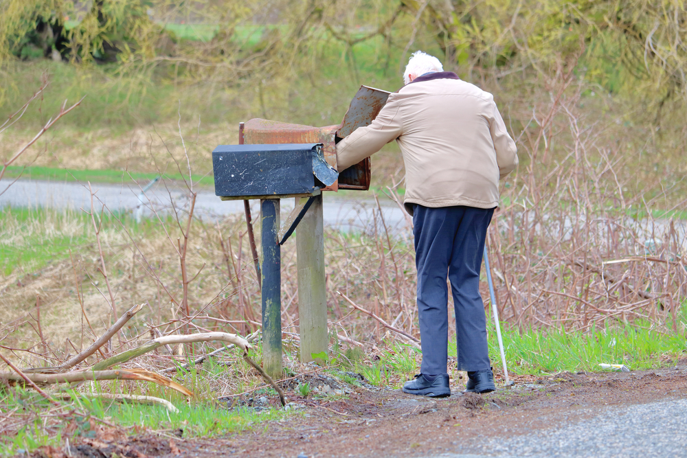 Old man with a white cane checks his mailbox for mail outdoors on a rural road.